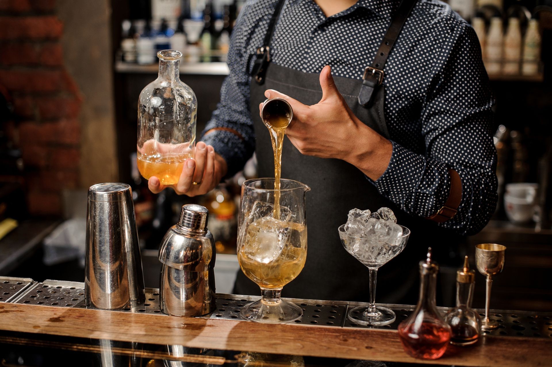 Barman pouring a portion of syrup into the large cocktail glass with ice cubes on the bar counter