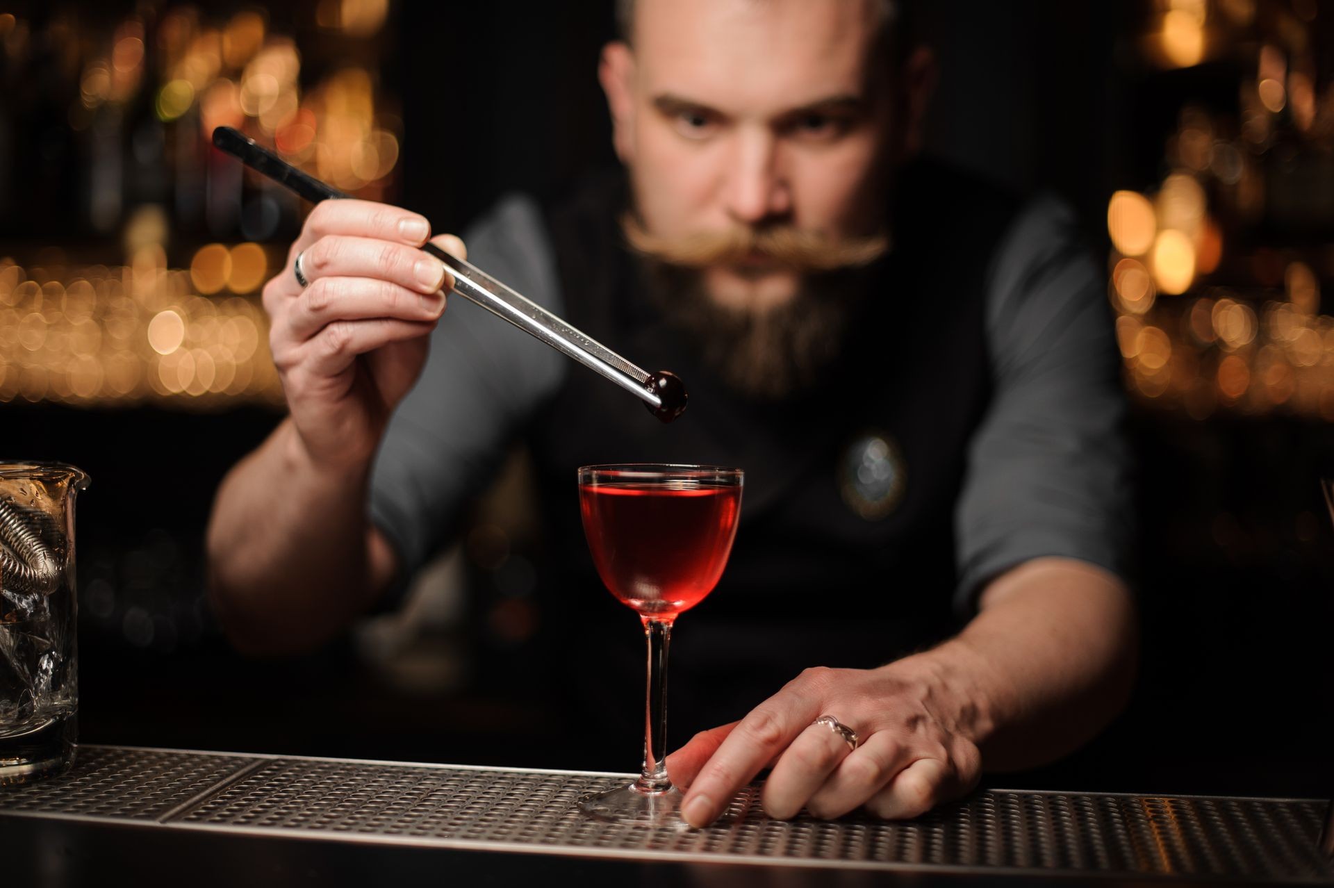 Portrait of male bartender with beard dropping small cherry from special tongs in an alcohol drink